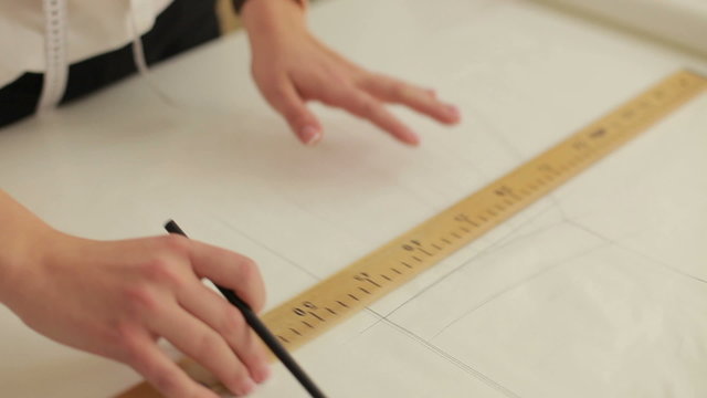 Fashion designer working on table. Hand of female tailor drawing pattern at paper in her studio. Young female tailor with measuring tape on shoulders working in workshop.