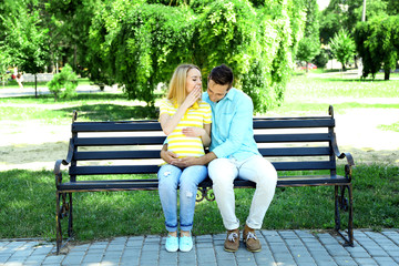 Young pregnant woman with husband sitting on bench in park