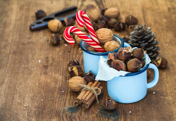 Traditional Christmas foods and decoration. Roasted chestnuts in blue  enamel mug, walnuts, cinnamon sticks, candy canes, pine cone on rustic wooden background