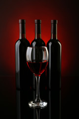 Wine glass against bottles in a row on dark red background, close up