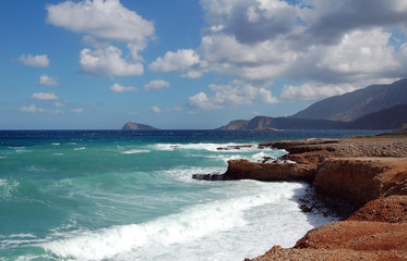 Waves on red rock sea shore with mountains and blue sky
