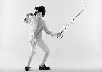 Boy wearing white fencing costume and black fencing mask standing with the sword practicing in...