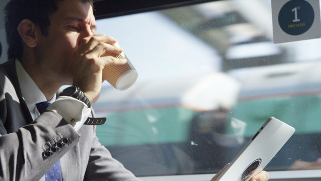  Businessman working on digital tablet & drinking coffee on train journey. Shot on RED Epic.