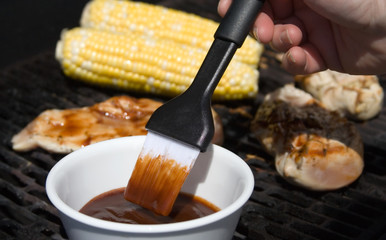 Barbecue Sauce and Food on the Grill – A brush with barbecue sauce on it in the foreground, corn on the cob and chicken on the grill in the background.