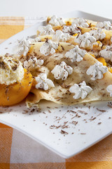 Crepes stuffed with vanilla cream and peach, decorated with whipped cream and grated chocolate, served on a white plate.