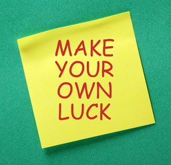 The message Make Your Own Luck in red text on a yellow sticky note posted on a green notice board as a reminder