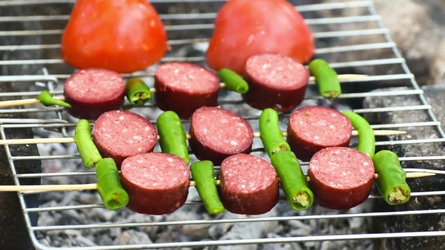 Turkish Sausage ( Sucuk ) cooking on the grill over coals. There are tomato and green pepper on the grill.
