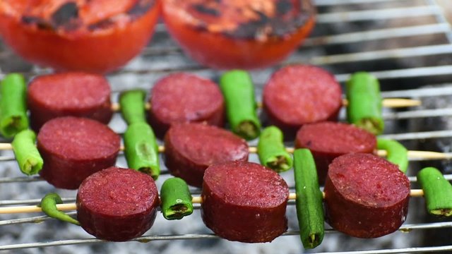 Turkish Sausage ( Sucuk ) cooking on the grill over coals. There are tomato and green pepper on the grill.
