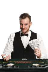 Mature casino worker wearing a black vest and white shirt with a bowtie. Holding a fan of dollar bills with a deck of cards spread in front of him. White background.