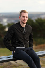 Handsome man wearing a black jacket and jeans sitting down outside on a bench on a cold windy...