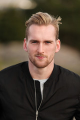 Handsome man wearing a black jacket and jeans sitting down outside on a bench on a cold windy autumn day. 