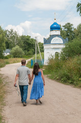 Obraz premium Father join hands with daughter in blue sarafan go on dirty road toward chapel. Dmitrievskoe village, Tulsky region, Russia. 