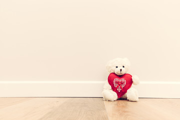 Cute teddy bear with big red plush heart. Sitting on wooden floor against white wall.