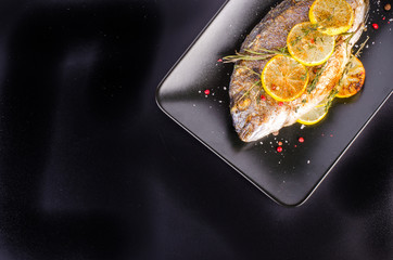 Roasted gilthead fishes with lemon, herbs, salt on black background. Healthy food concept. Food frame. Free space for your text.