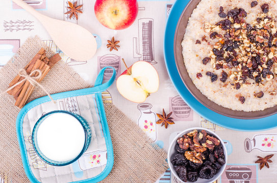 Breakfast set. A bowl of barley porridge with cocnut oil, vegetarian milk, walnuts, raisins, cinnamon on sackcloth, canvas, spoon, apple, glass of vegetarian milk.