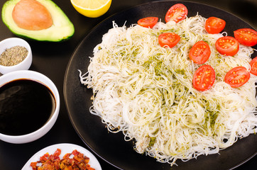 Chinese rice noodles with pesto, tomatoes cherry, avocado, soy sauce, lemon and spices on black background.