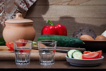 Ужин в деревне. На столе овощи, две рюмки, кастрюля с едой. Хлеб в тарелке и яйца. Яблоки.  Овощи - помидоры, огурцы, перец. Овощи нарезанные и целые. Фон - деревянная доска. Алкоголь