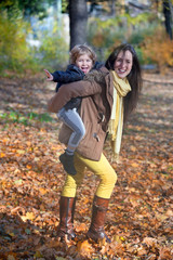 Cheerful mother giving her son piggyback ride and having fun with him in park in autumn.