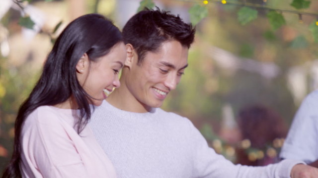 Attractive Asian couple cooking at bbq while friends socialize in background