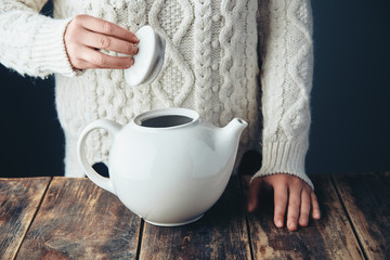 Woman in warm sweater holds cap of big white teapot in hands