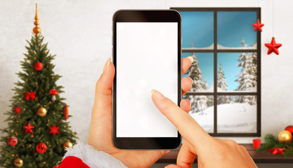 Woman with smart phone in hands in Christmas time. Device with white screen for mockup. Christmas tree, gifts, decorations in background.