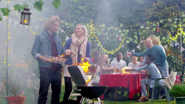 Happy attractive couple cooking at bbq while friends socialize in the background
