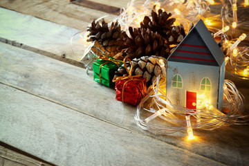 Fototapeta na wymiar pine cones and decorative wooden house next to gold garland lights on wooden background. copy space 