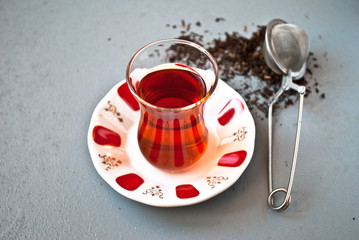Turkish tea in traditional glass and tea strainer in a pile of tea leaves on a light gray background