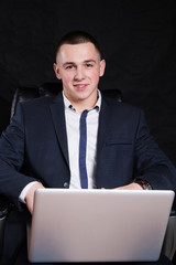 Successful young businessman in a blue suit sits in a chair and holding a laptop.