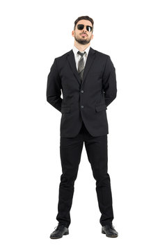 Secret agent or guard with hands behind back wearing sunglasses.Full body length portrait isolated over white studio background.