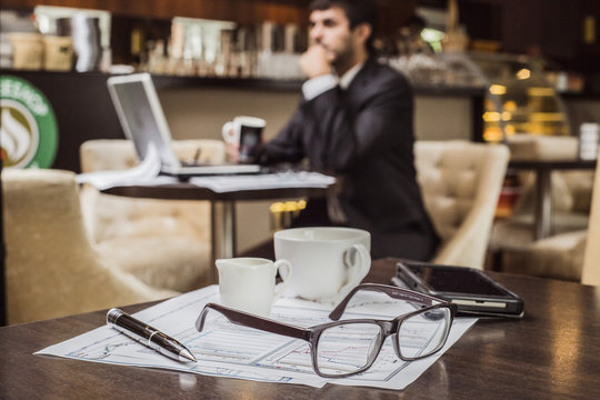 Бизнесмен работает в кафе, а перед ним на столе лежат атрибуты делового человека - очки, ручка и деловые бумаги. 