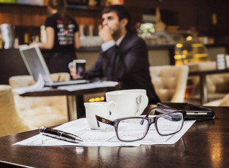 На столе лежат атрибуты бизнесмена и инвестора, финансовые бумаги, графики и   очки, за ними на размытом фоне силуэт бизнесмена фрилансер