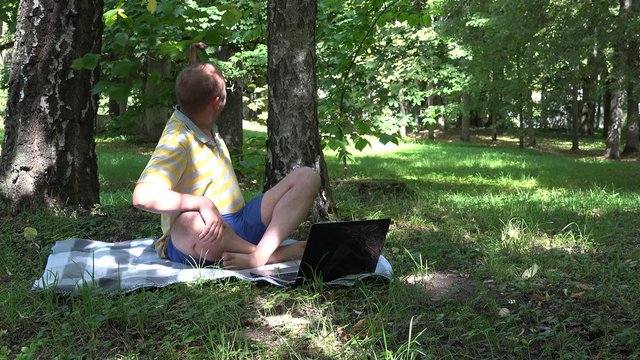 Relaxed man with laptop communicating online with girlfriend on his holiday in park. Static shot. 4K