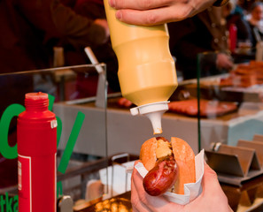 Hand pressing a bottle with mustard on a hot dog with a german sausage in a paper napkin