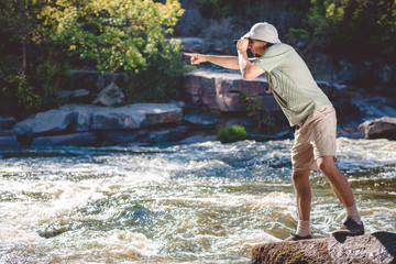 Man in pith helmet standing on rocky riverbank using binoculars 