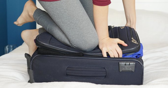 Young woman standing on her knees on overfilled suitcase, trying to close it