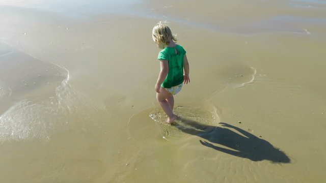 tender funny scene of two years age blonde baby with green shirt and swimwear shorts jumping water puddle and playing on golden sand beach seaside