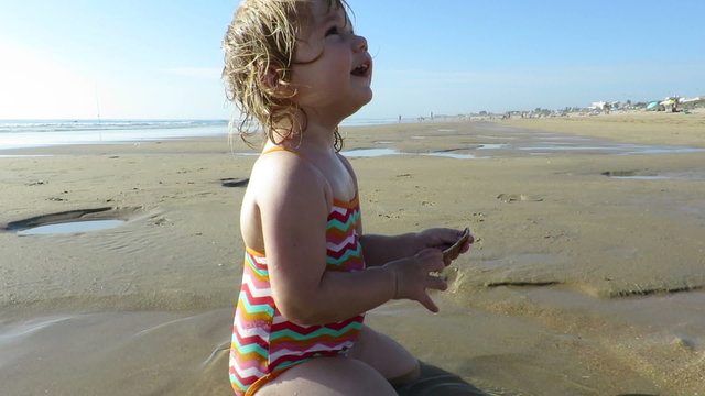 two years blonde baby swimsuit colored stripes looking for, picking and collecting sea shells in puddle in golden sand beach seaside next to woman mother or babysitter