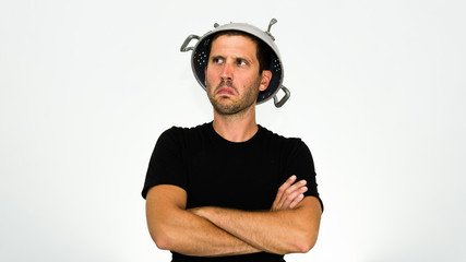 close-up of angry young caucasian man with a colander on his head and arms crossed looking at his...
