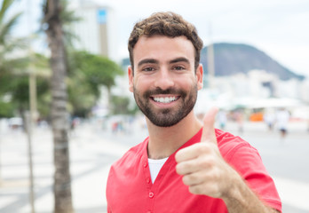 Junger Mann mit Bart und rotem Shirt in der Stadt zeigt Daumen
