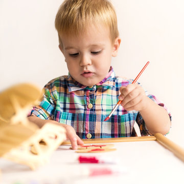 Adorable kid playing on his table with watercolors, painting a wooden parts of his airplane wooden toy. He is young designer with many ideas waiting to be found. Very shallow depth of field.