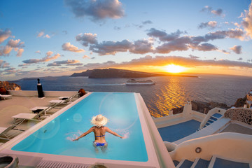 Obraz premium Woman in the swimming pool against sunset in Oia village on Santorini island, Greece