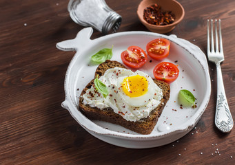Sandwich with feta cheese and boiled egg, tomatoes, and basil on a white plate on a dark wooden surface. Healthy breakfast or snack