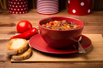 Chili con carne served in the red bowl on the wooden background.