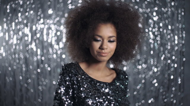 Young African American woman posing in an elegant sparkling black party dress against modern silver background inside a studio.