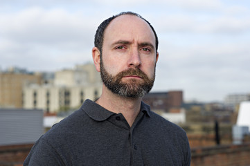 Portrait of man on a London rooftop. He has a balding head and neatly trimmed beard. 