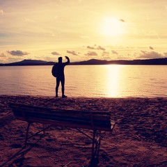 Alone traveler with backpack. Man  on sea beach at wooden bench, cold sunny autumn evening