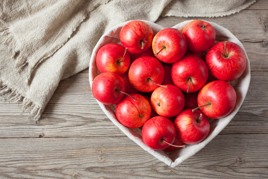  Red apples in a basket in the shape of a heart on a wooden background.