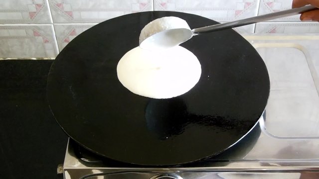 Dosa batter is being poured, in a circular way, on a hot skillet.