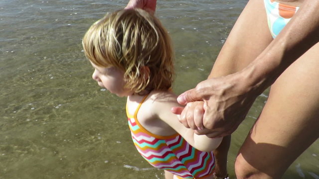 slow motion tender family  two years age blonde baby with swimsuit colored stripes with mother bikini woman walking, running and playing water waves in beach seaside ocean